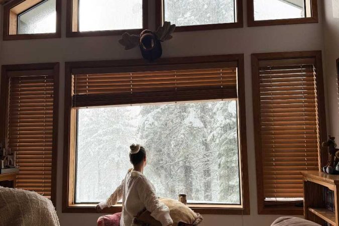 Femme assise près d'une fenêtre à double vitrage avec stores en bois, regardant un paysage enneigé depuis l'intérieur d'une maison bien isolée.