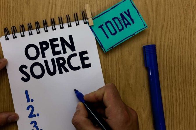 Une main écrit sur un carnet de notes avec l'inscription Open Source à côté d'un marqueur bleu sur une table en bois.