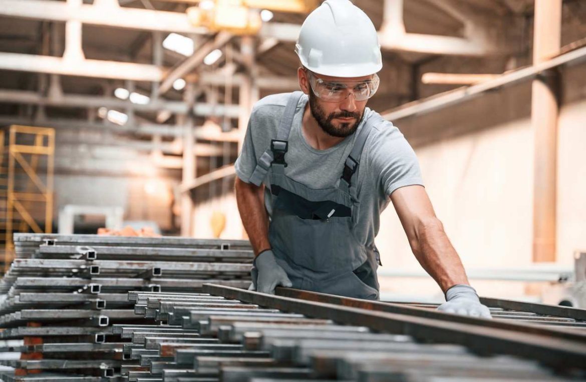 Ouvrier en casquet de chantier manipulant une barre de fer sur un stock de profilés en acier dans un atelier.