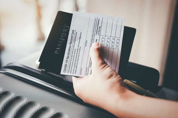 Gros plan sur une main tenant un passeport et une carte de vaccination COVID-19 près d'une valise, illustrant des documents de voyage essentiels.