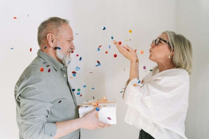 Un couple de seniors célèbre un heureux anniversaire de mariage en soufflant des confettis colorés et en tenant un cadeau.
