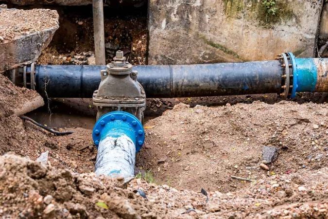 Une tranchée creusée dans la terre expose un système de tuyauterie souterrain composé de gros tuyaux noirs et d'un tuyau en PVC fêlé à la jonction avec une bride bleue.