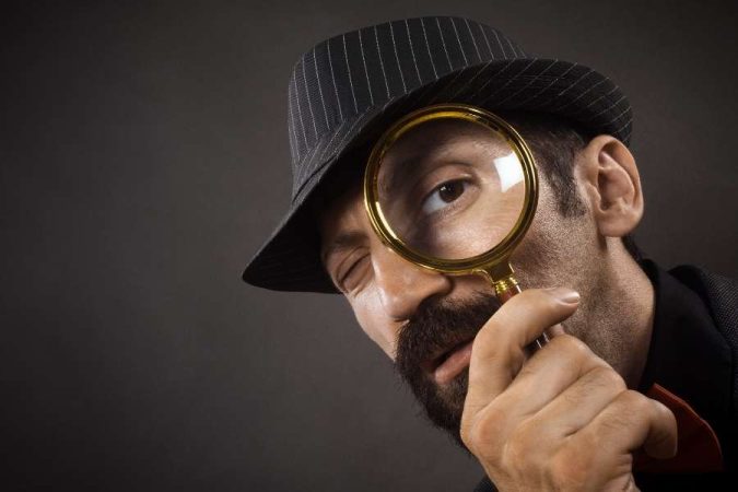 Un détective privé barbu en chapeau fedora observe attentivement à travers une loupe.
